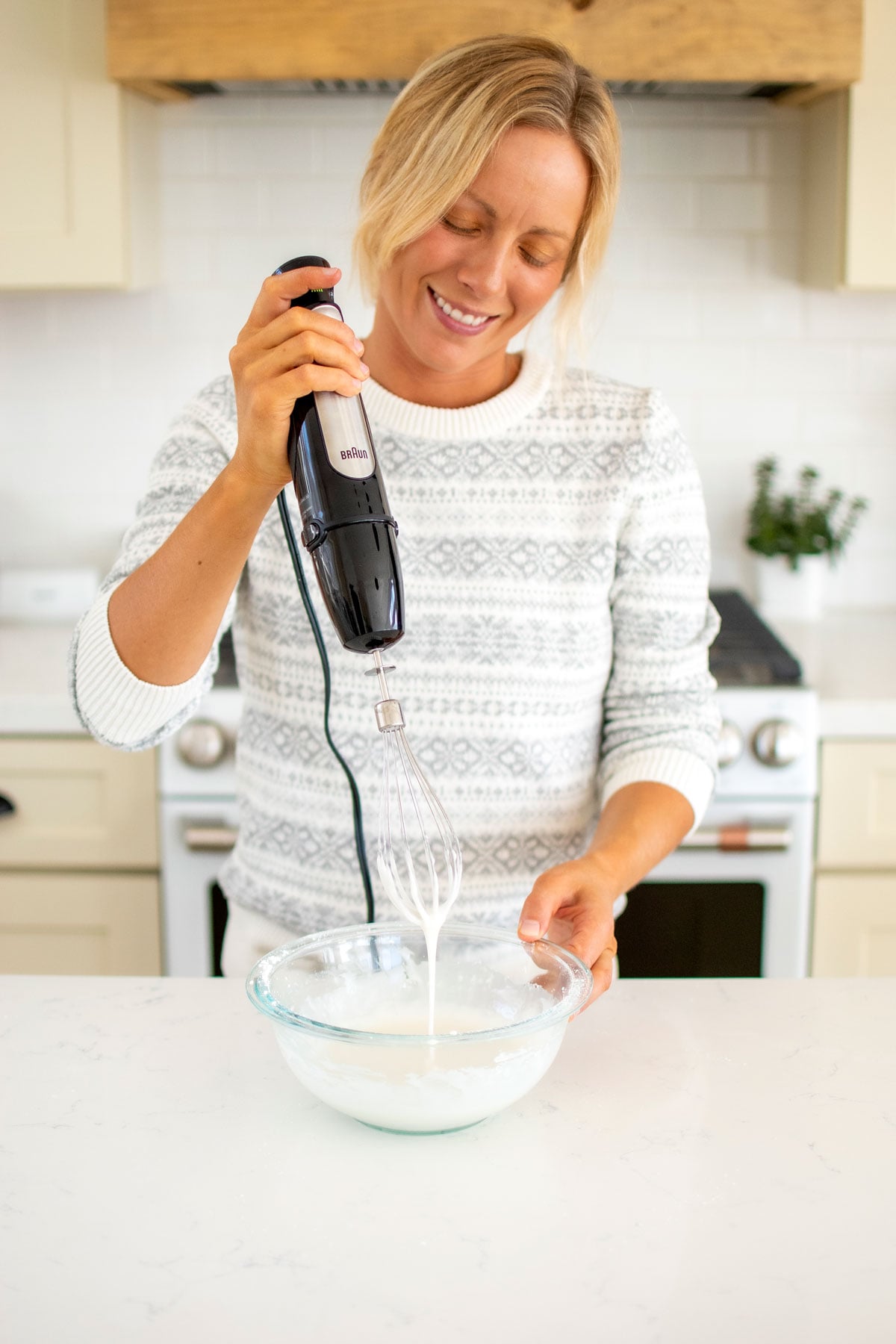 woman mixing glaze in a glass bowl at the kitchen counter using a hand mixer.