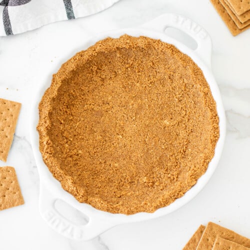 the best homemade graham cracker crust in a white pie dish on a marble counter with graham crackers around it.