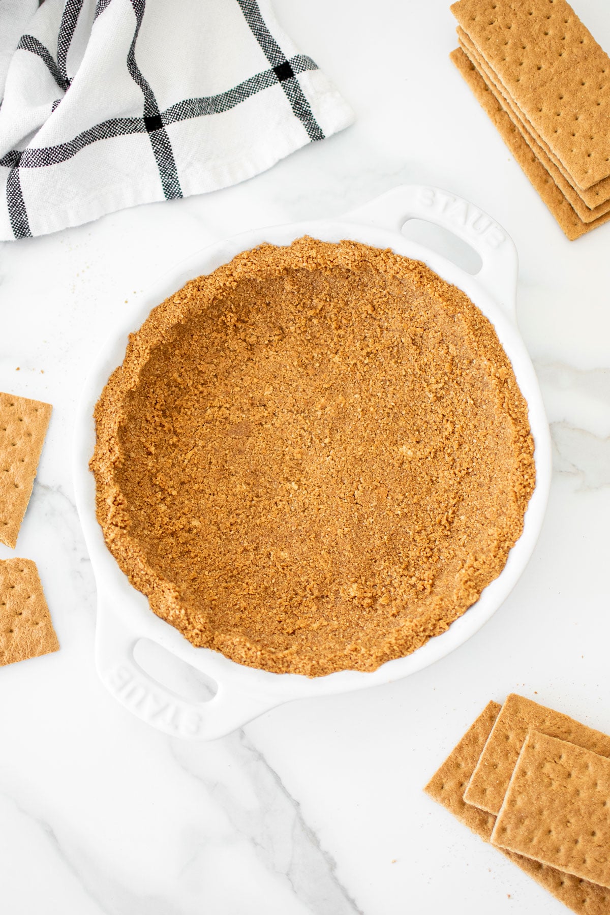 the best homemade graham cracker crust in a white pie dish on a marble counter with graham crackers around it.