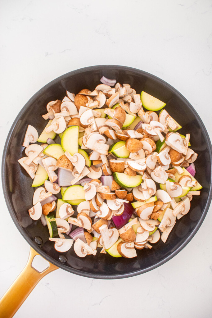 sliced mushrooms, zucchini and red onion in a large skillet on a marble countertop.