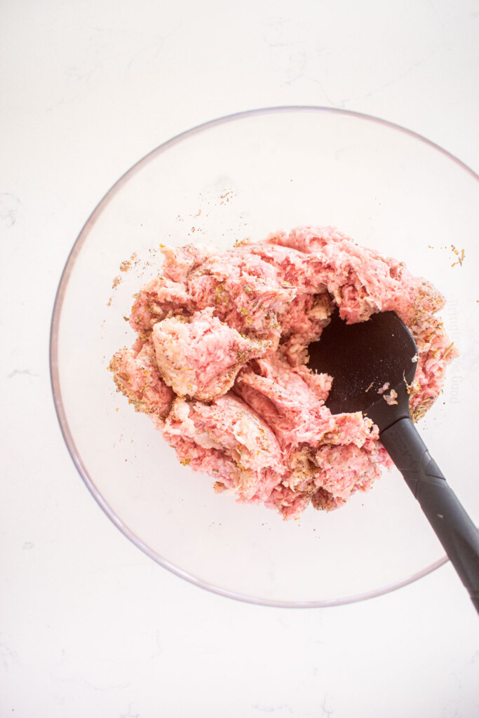 raw ground beef with spices in a glass mixing bowl on a marble countertop with a spatula in it.
