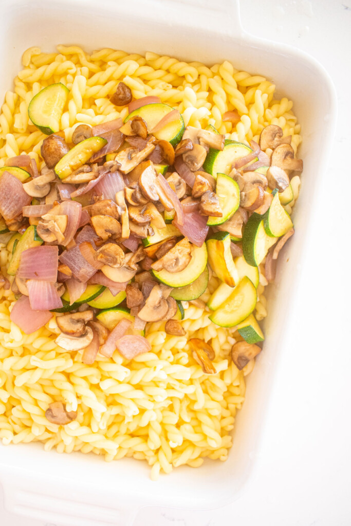cooked noodles, zucchini, mushroom and onions in a white baking dish on a marble countertop.