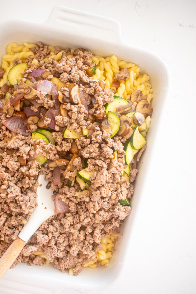 cooked noodles, zucchini, mushroom onions and ground beef in a white baking dish on a marble countertop.