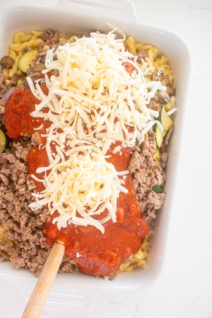 cooked noodles, zucchini, mushroom onions, ground beef, sauce and mozzarella in a white baking dish on a marble countertop.