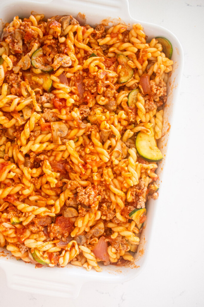 pasta with sauce, ground beef and veggies mixed together in a white ceramic dish on a marble countertop.