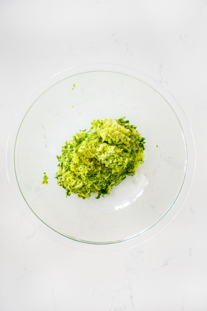 organi fine grated zucchini in a glass bowl on a white marble countertop.