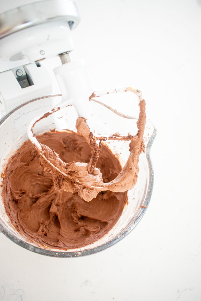 chocolate frosting in a glass mixing bowl on a white marble countertop.