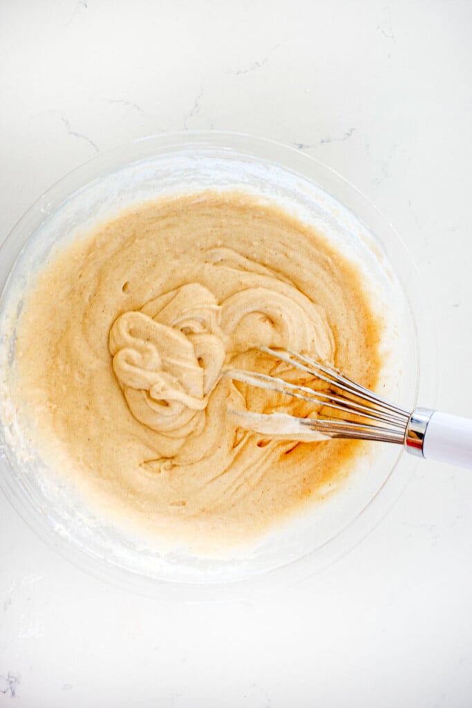 ingredients for sparkling apple cider donuts in a glass mixing bowl with a whisk on a white marble counter.