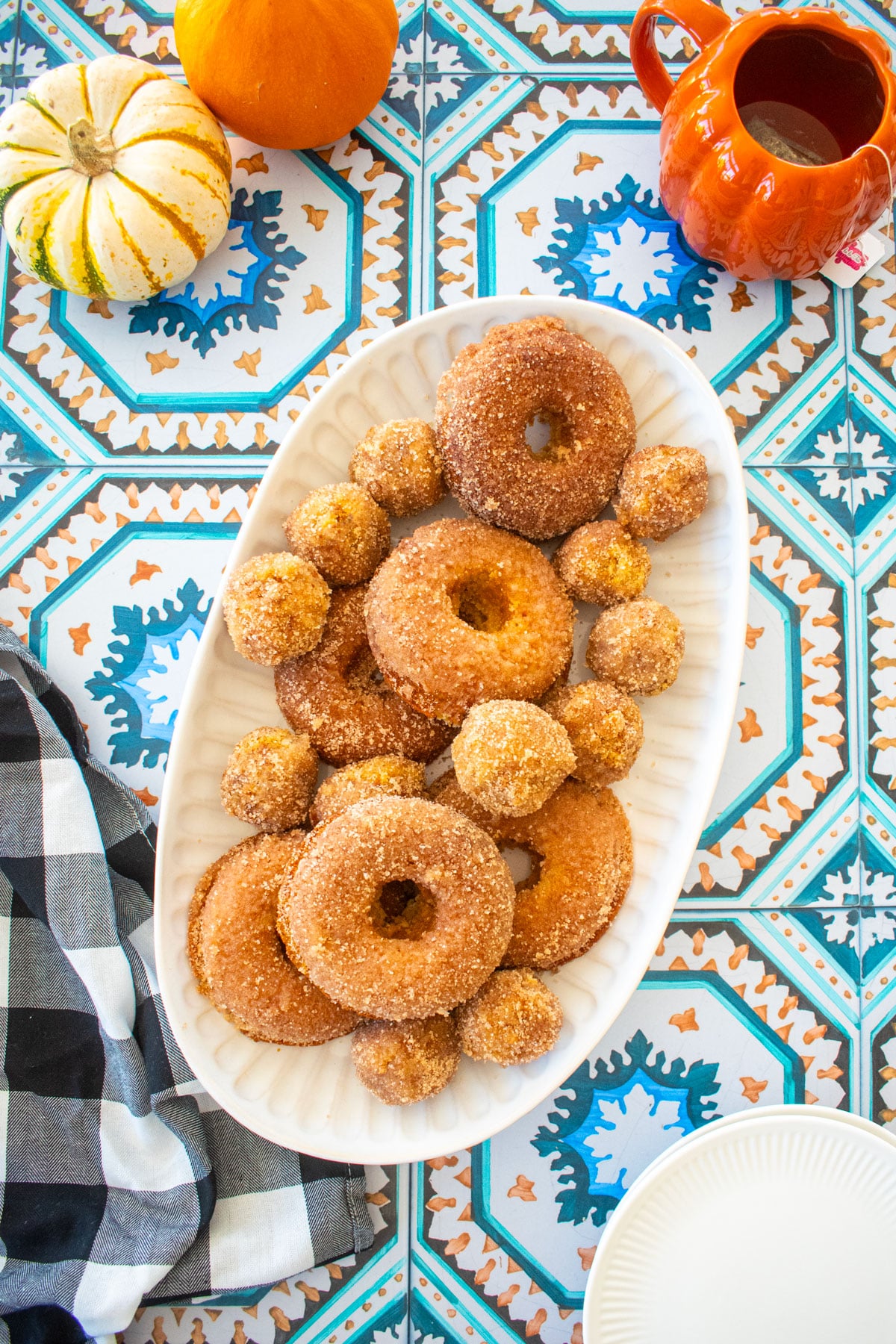 platter of sparkling apple cider donuts on a tile table with tiny pumpkins and a mug of tea.