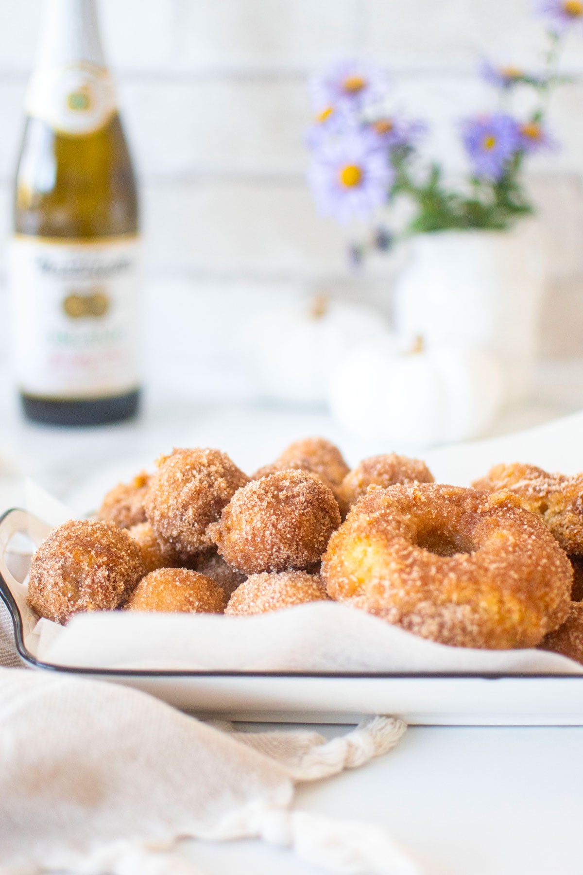 sparkling apple cider donuts on a white serving platter on a marble counter with fresh flowers.