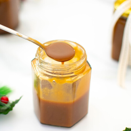 glass jar of brown butter butterscotch sauce with a spoon in it on a white marble counter.