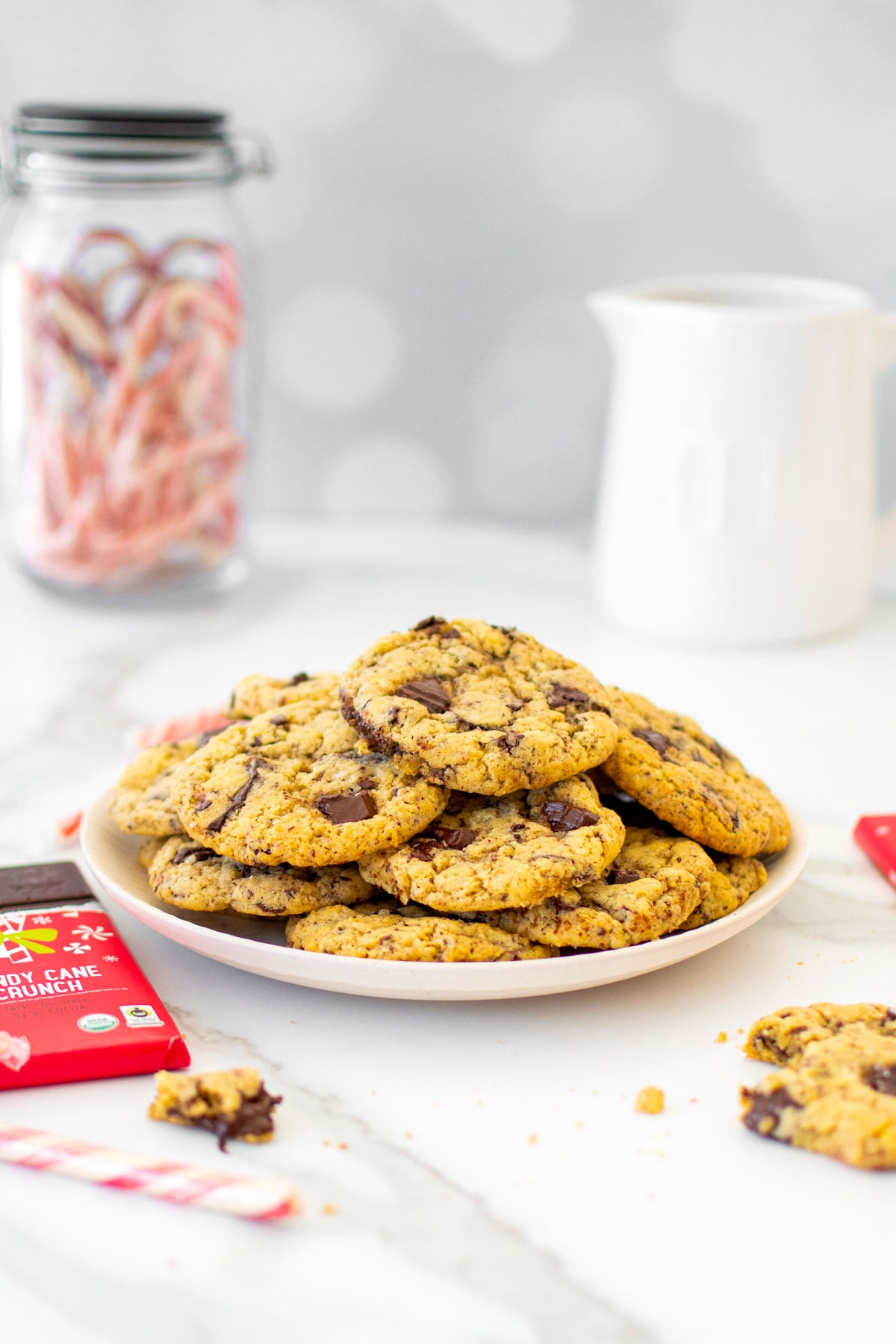 plate of candy cane crunch dark chocolate cookies on a white marble counter with a pitcher of milk and jar of candy canes.