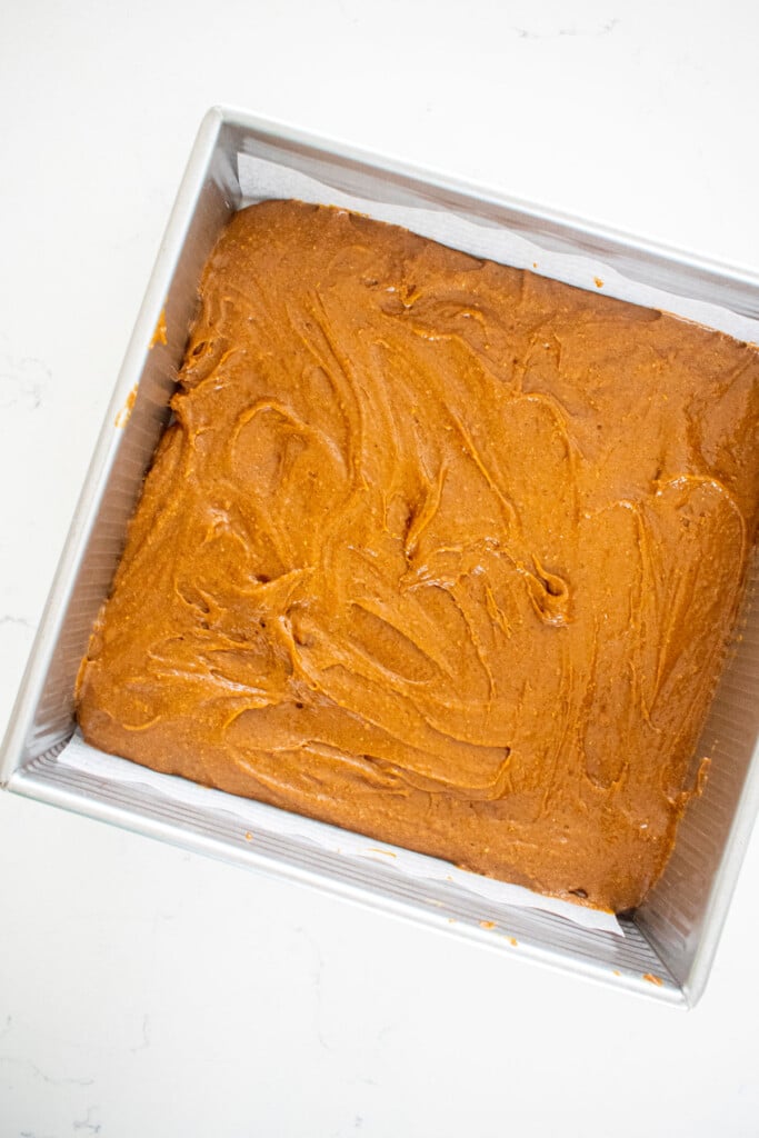 gingerbread cake batter spread into a square cake pan on a white marble counter.