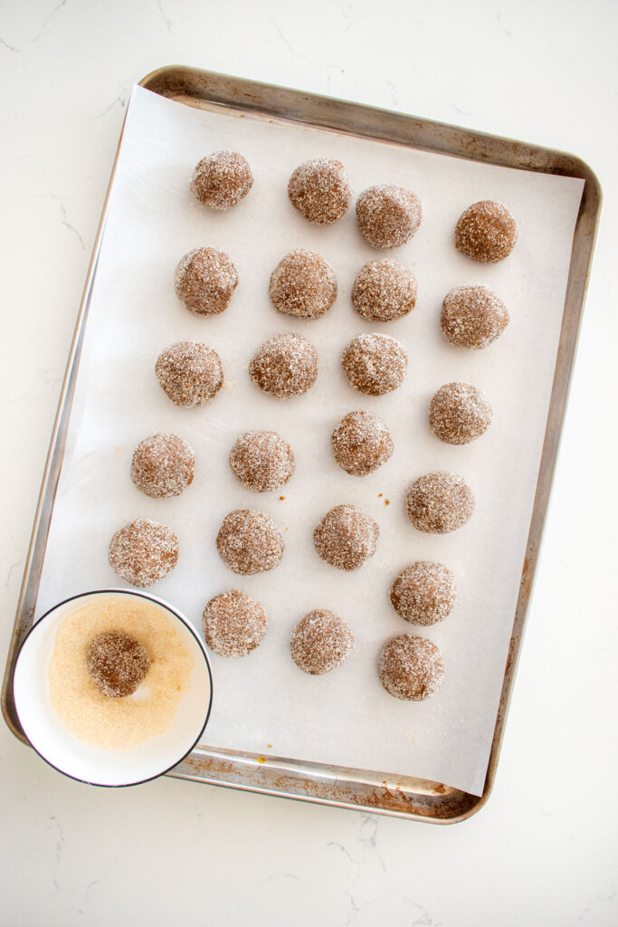 eggless sourdough molasses cookie dough balls rolled in cane sugar on a baking sheet on a white marble counter.