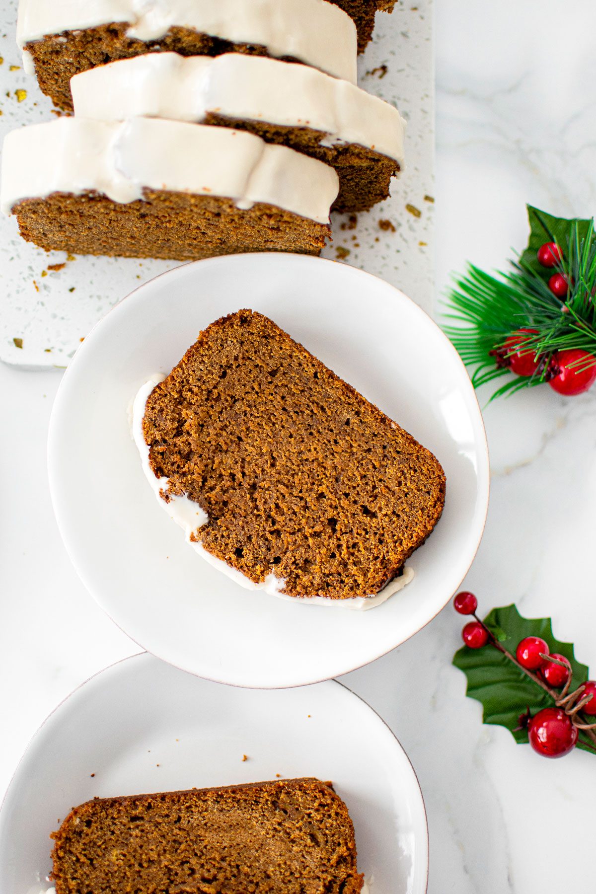 slices of frosted gingerbread banana bread on small white dessert plates on a marble counter with holly.