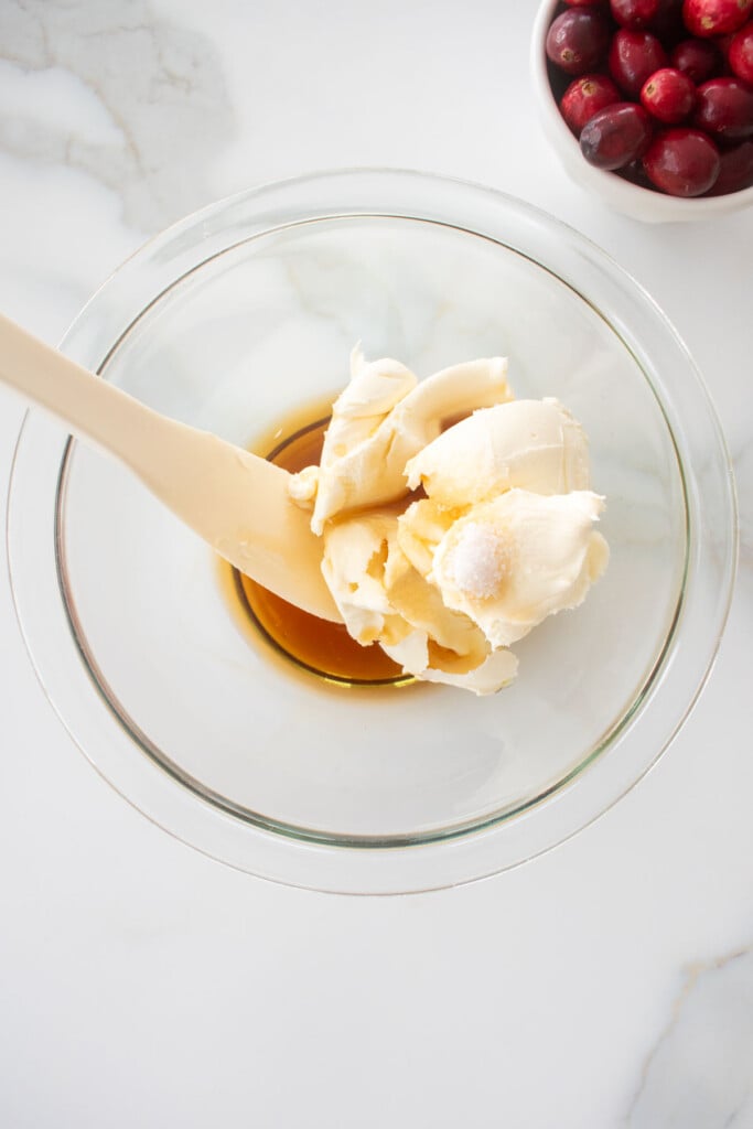 a glass bowl with cream cheese, maple syrup and salt on a white marble counter with a spatula in it.