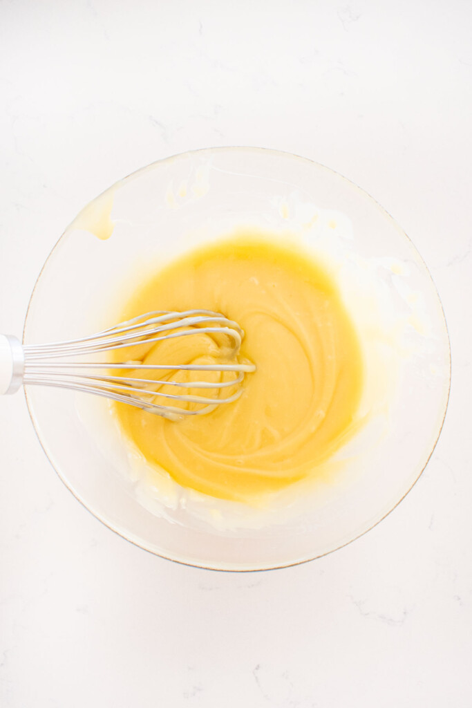 white chocolate ganache in a glass mixing bowl with a whisk on a white marble counter.