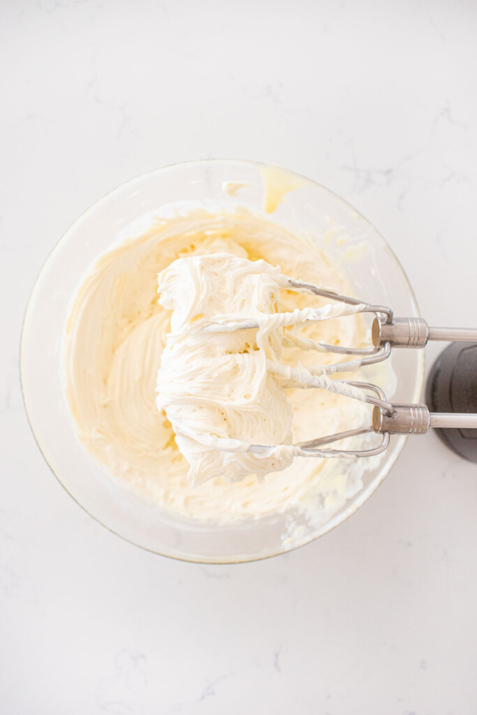 whipped white chocolate ganache in a glass mixing bowl with a hand mixer on a white marble counter.