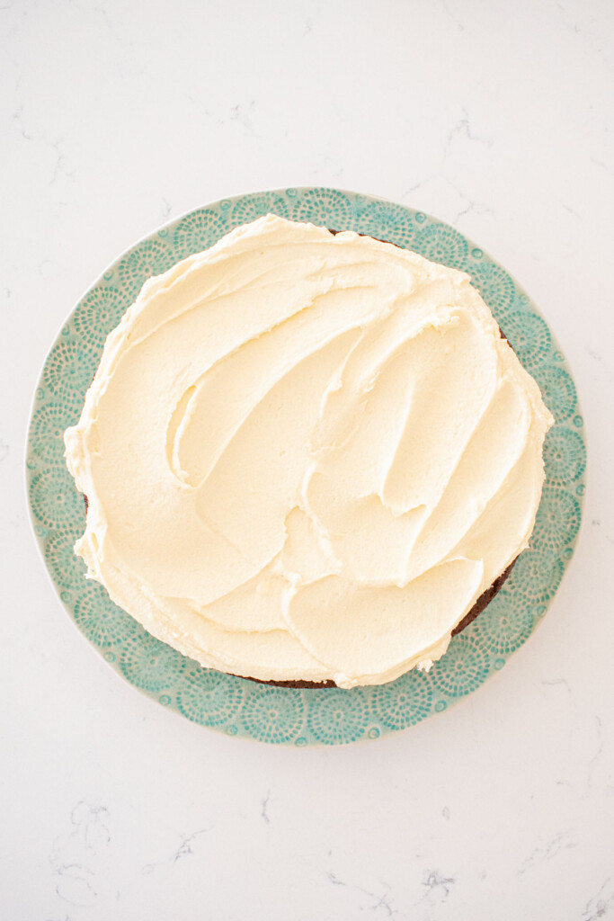 white chocolate ganache frosting on top of a chocolate cake on a green cake plate on a white marble counter.