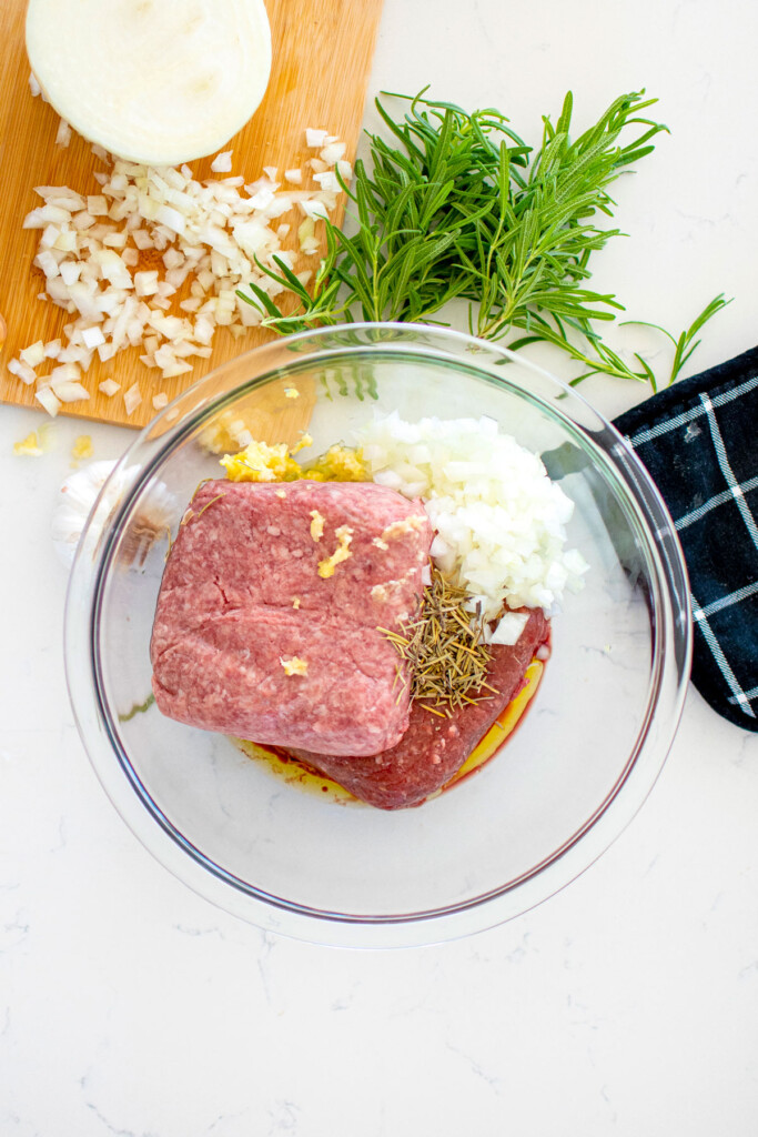 ingredients for rosemary meatballs in a glass mixing bowl on a marble countertop.