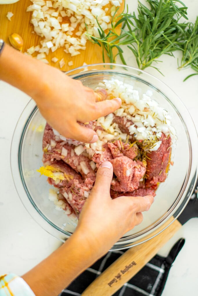 woman mixing rosemary meatball ingredients in a glass mixing bowl on a marble countertop.
