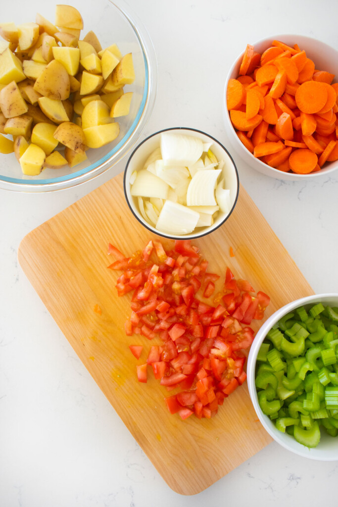 chopped potatoes, carrots, celery, onions in bowls and chopped tomatoes on a wood cutting board on a marble counter.
