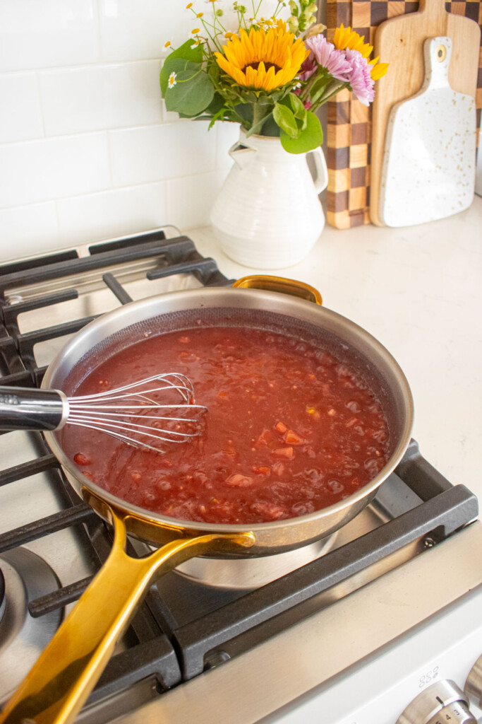 large skillet with beef broth, red wine, garlic, and tomato on a stovetop.