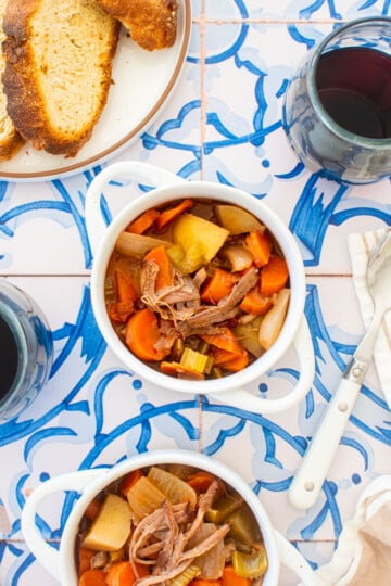 two mini dutch ovens with slow cooker beef brisket stew, stemless wine glasses and a bread plate on a blue tile table.