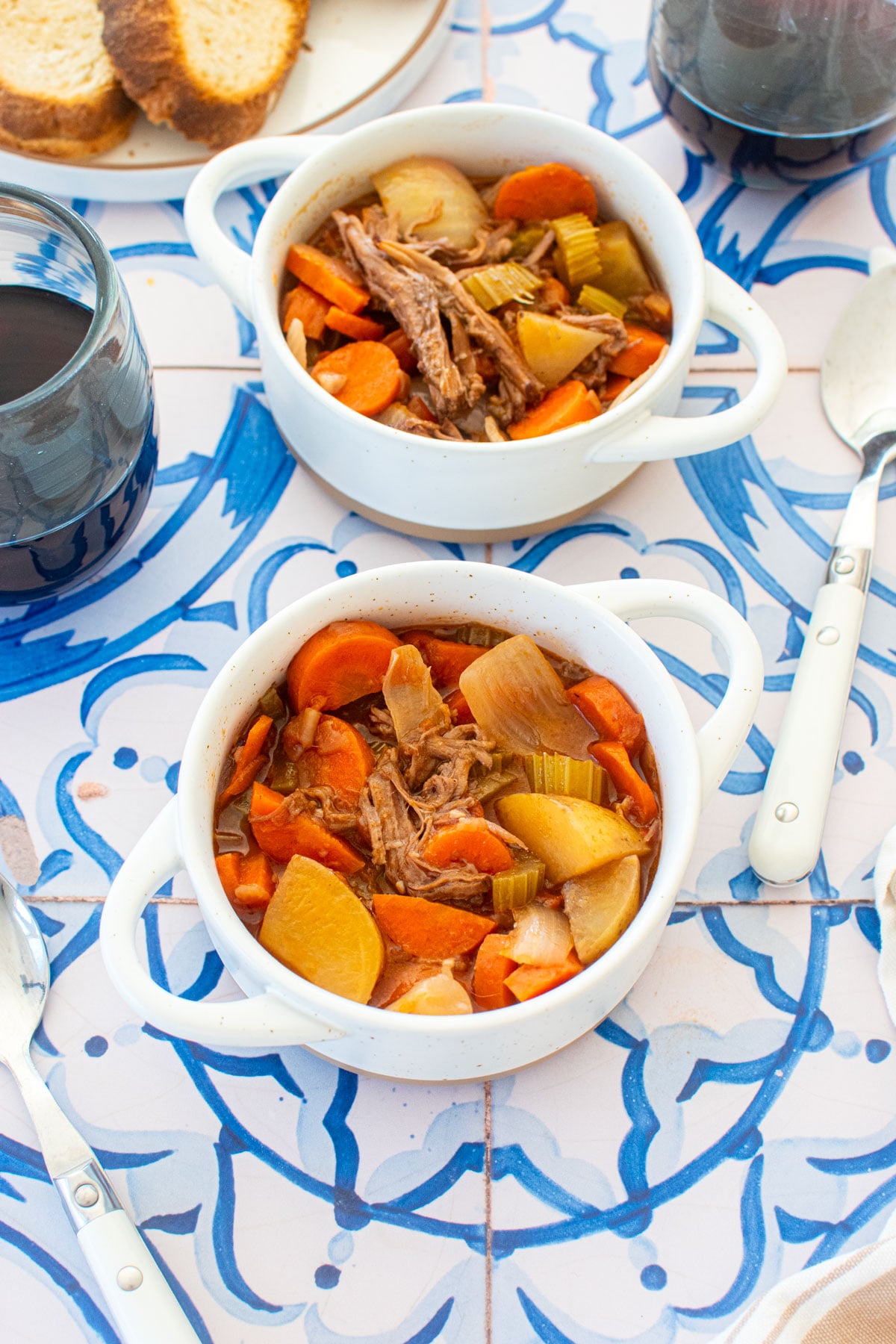 two mini dutch ovens with slow cooker beef brisket stew, stemless wine glasses and a bread plate on a blue tile table.