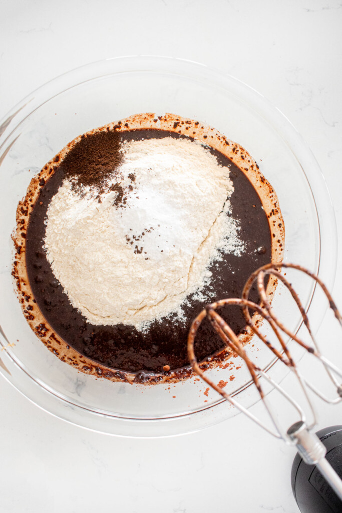wet ingredients for vegan brownies in a glass mixing bowl with dry ingredients added on a marble counter.
