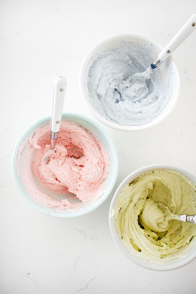 three bowls with light pink, light blue, and light green frosting on a white marble counter.