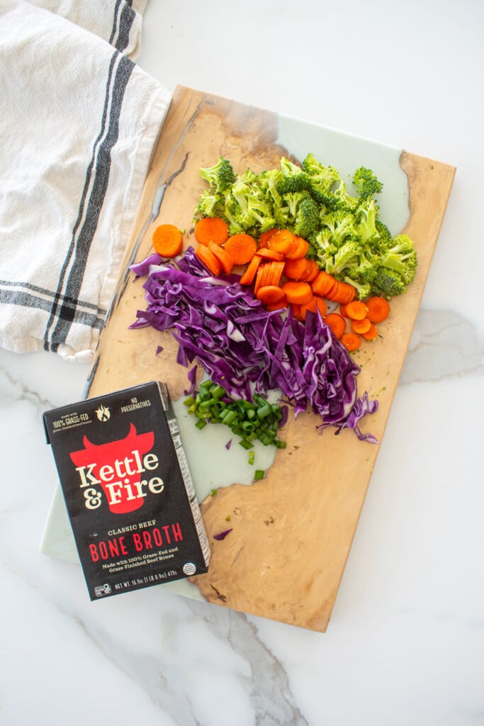 wood cutting board with chopped broccoli, carrots, purple cabbage and green onion with a carton of kettle and fire beef bone broth on a marble counter.