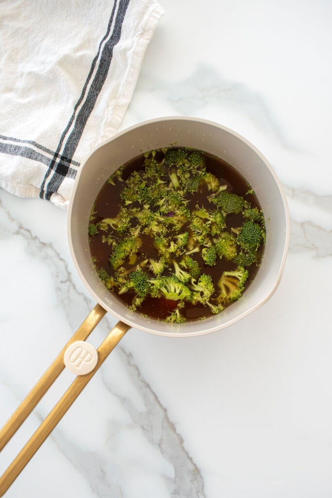small pot with bone broth, broccoli and carrot on a marble counter.