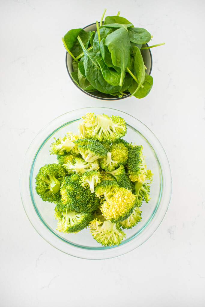 bowl of broccoli florets and a bowl of spinach on a white marble counter.