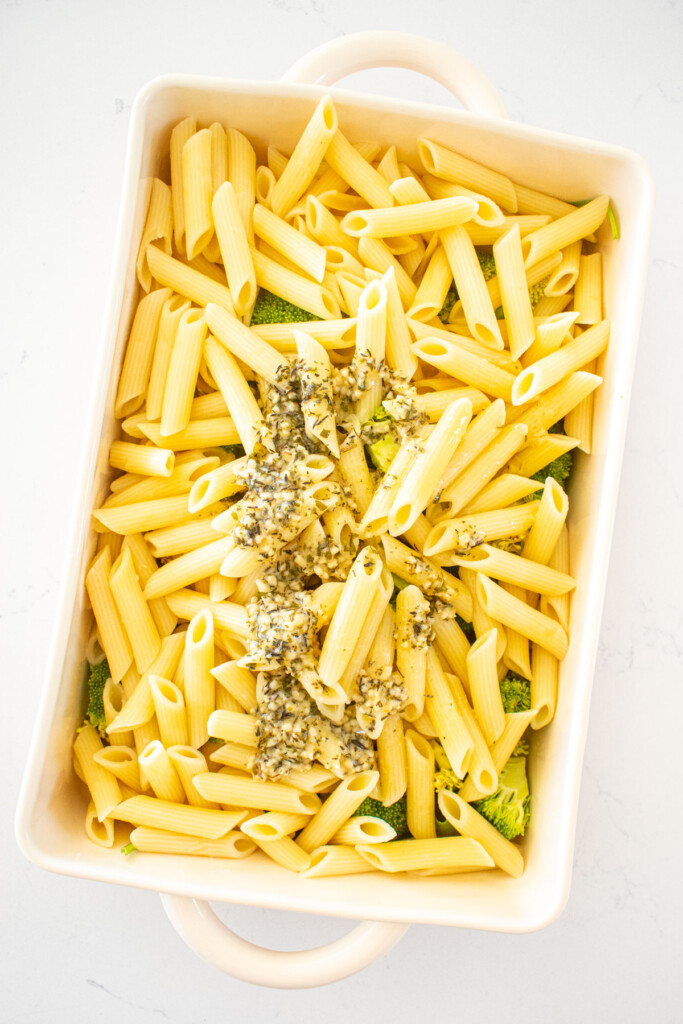 baking dish on a white marble counter with broccoli, spinach, penne pasta and herb butter.