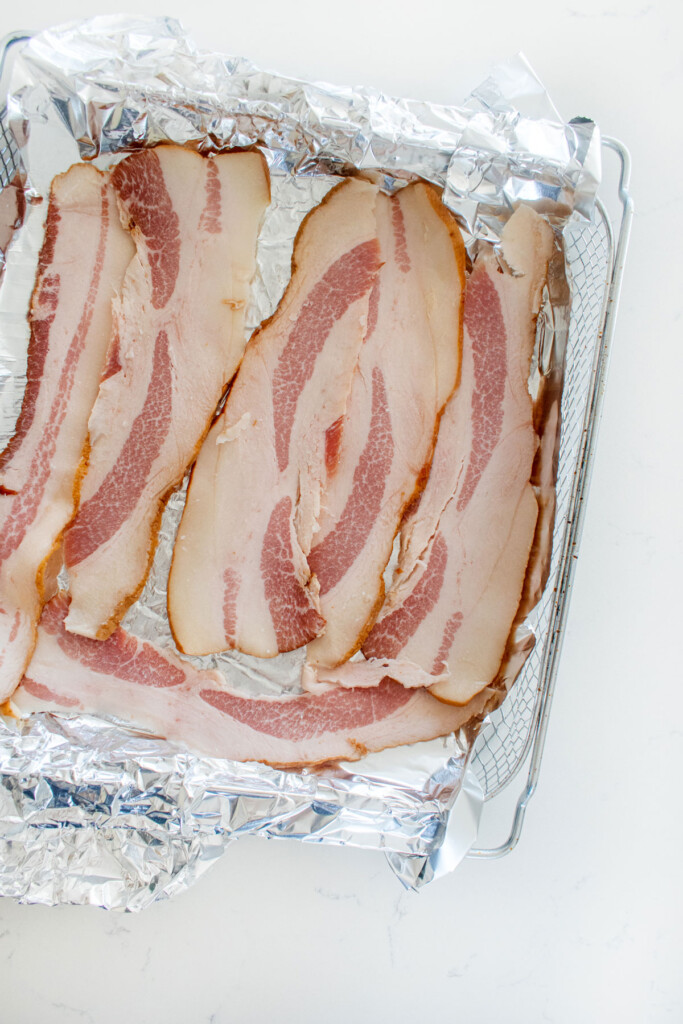 raw bacon on foil in an air fryer basket on a white marble countertop.