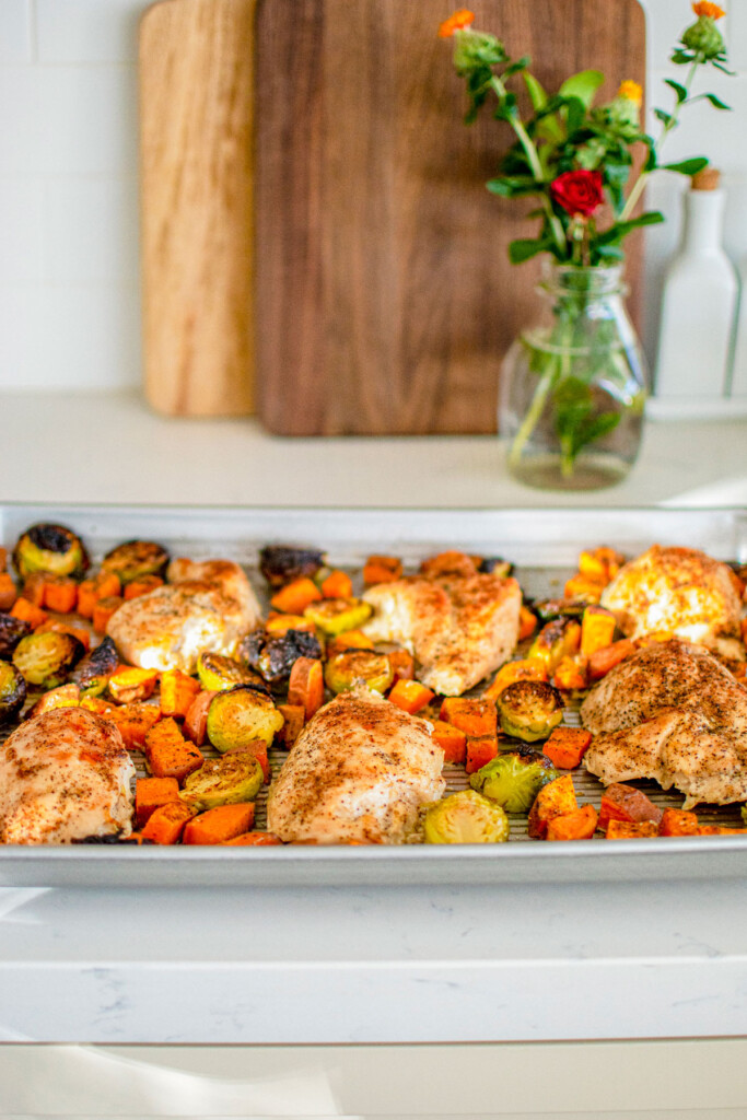 cooked chicken breasts on a sheet pan with charred brussels sprouts, sweet potato and bacon bits on the kitchen counter.