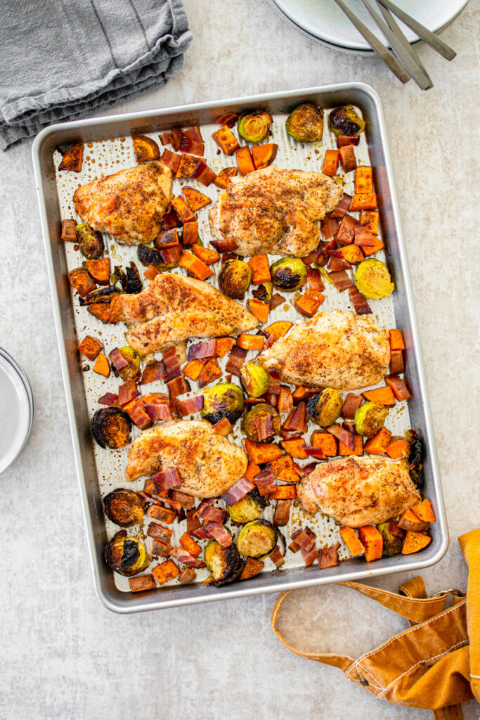 cooked chicken breasts on a sheet pan with charred brussels sprouts, sweet potato and bacon bits.