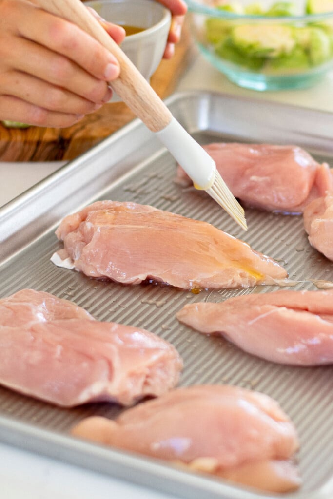 woman brushing ghee onto raw chicken breasts on a baking sheet.
