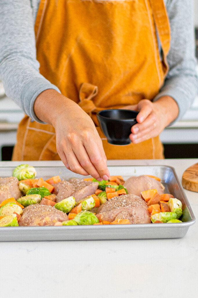 woman sprinkle seasoning mix on top of raw chicken breasts, sweet potato and brussels sprouts on a baking shseet.