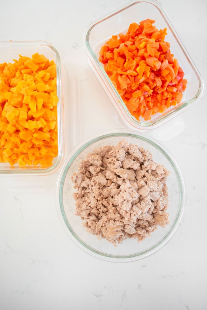 ground chicken, sweet potatoes, bell pepper and carrots in glass tupperware on a white marble countertop.