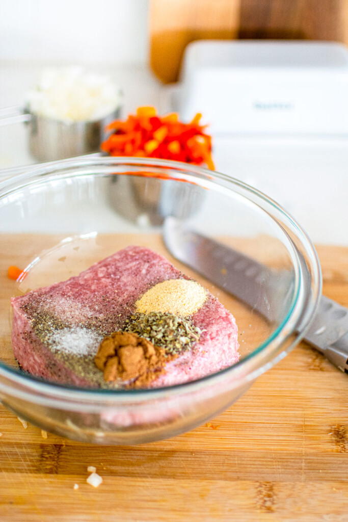 raw ground pork in a glass mixing bowl with spices on a wood cutting board on the counter.
