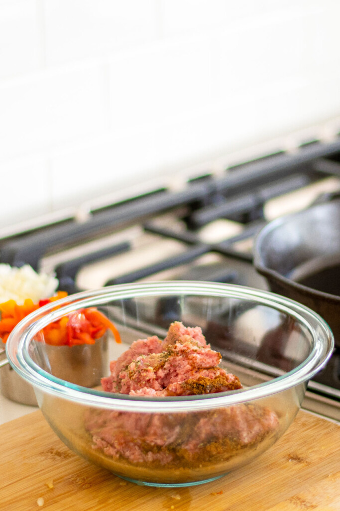 raw ground pork in a glass mixing bowl mixed together with spices on a wood cutting board on the counter.