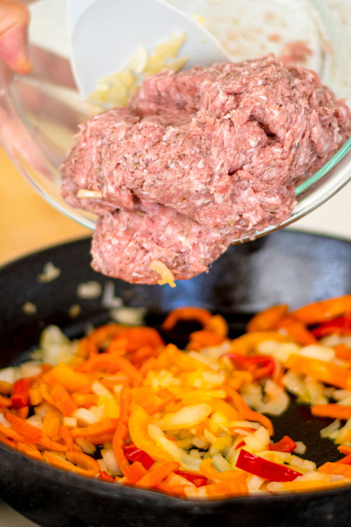 ground pork being added into a skillet with peppers and onions on the stove.