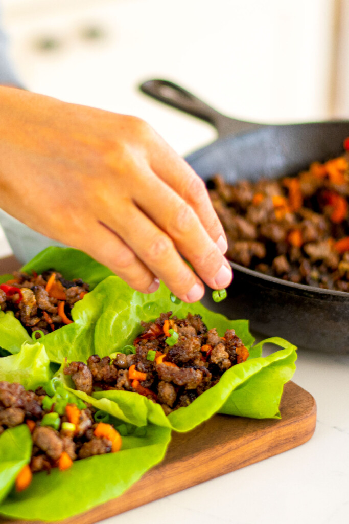 woman putting green onions on top of pork lettuce cups on a wood serving board.