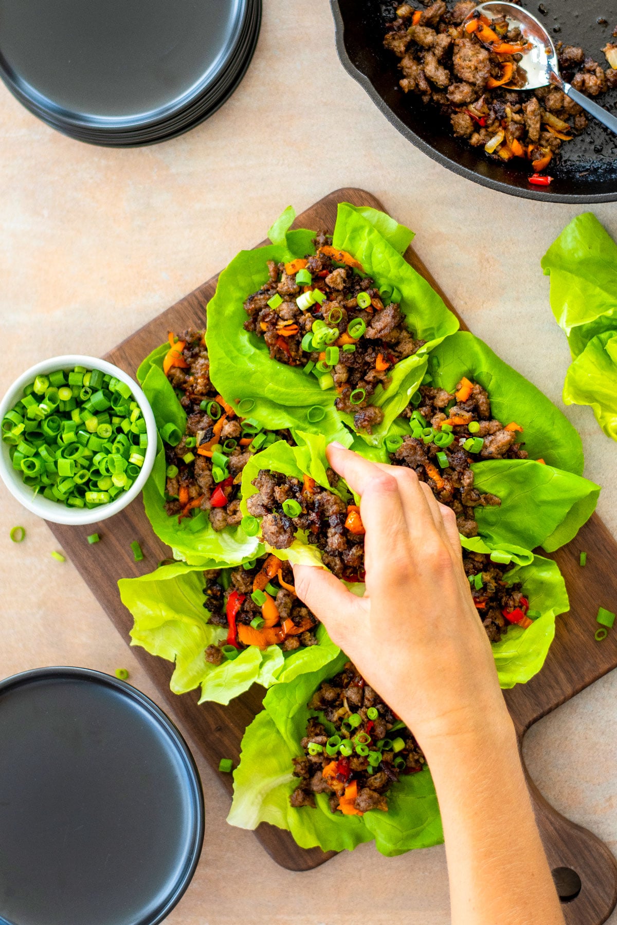 chinese five spice lettuce cups with pork on a wood serving board with a small bowl of green onions, woman grabbing one.