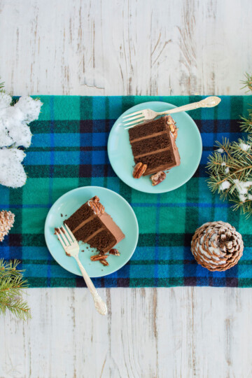 chocolate cake slices on a green plates on a white wood table with a plaid runner and pine cones.