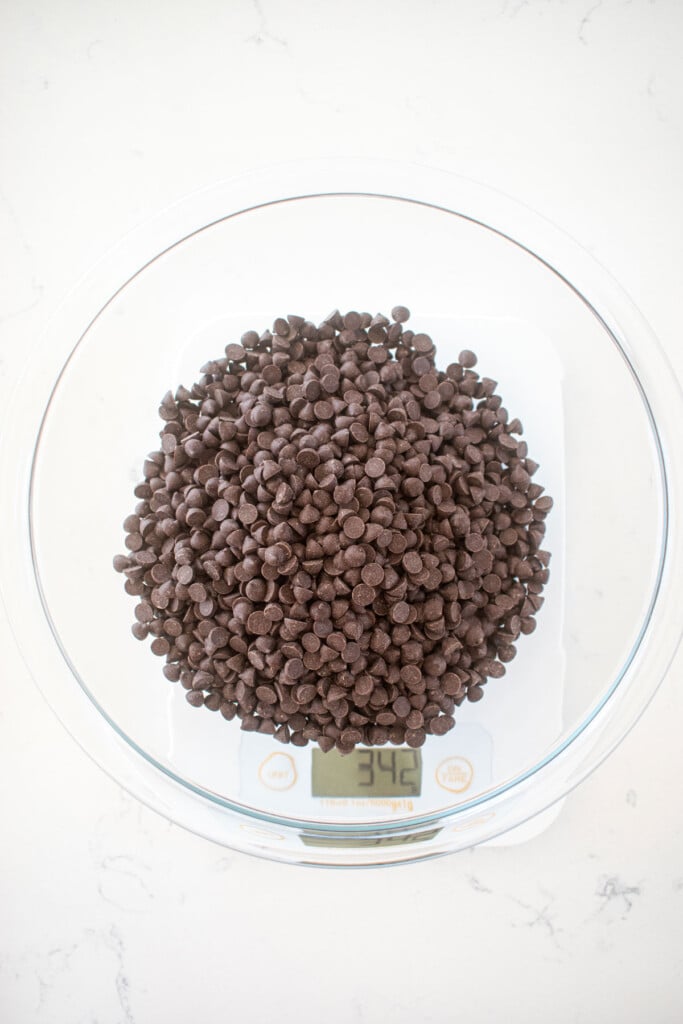mini dark chocolate chips in a glass mixing bowl on a scale on a marble counter.