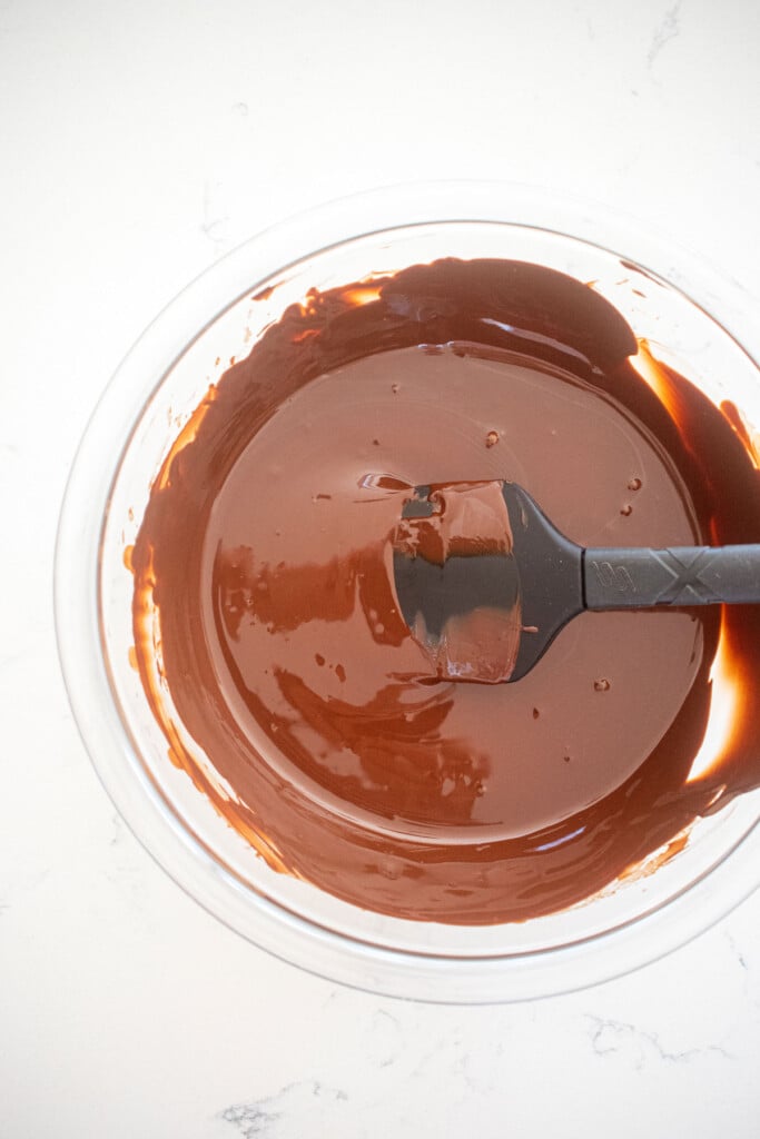melted dark chocolate in a glass mixing bowl with a spatula on a white marble counter.