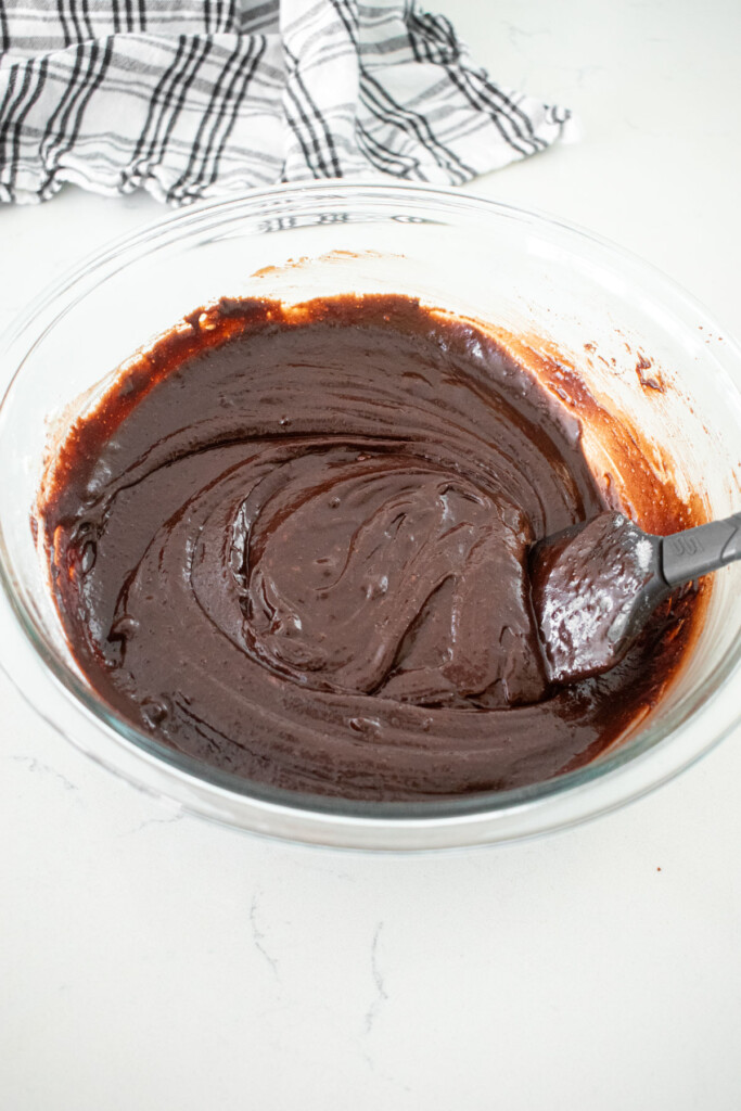 smooth brownie batter in a glass mixing bowl with a spatula on a white marble counter.